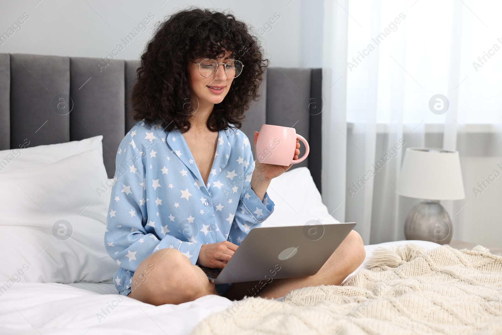Photo of Beautiful young woman in stylish pyjama with cup of drink using laptop on bed at home