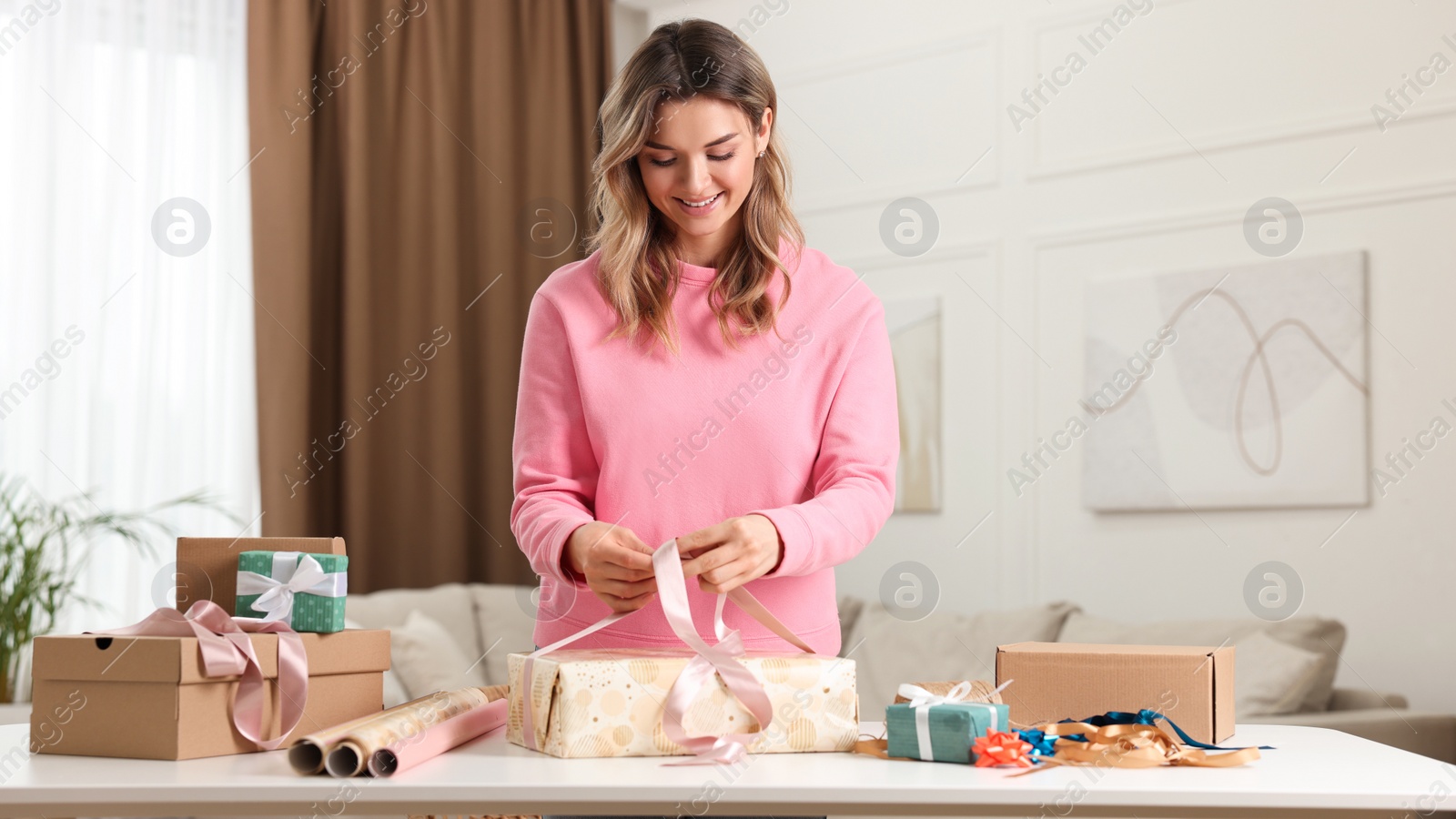 Photo of Beautiful young woman wrapping gift at table in living room