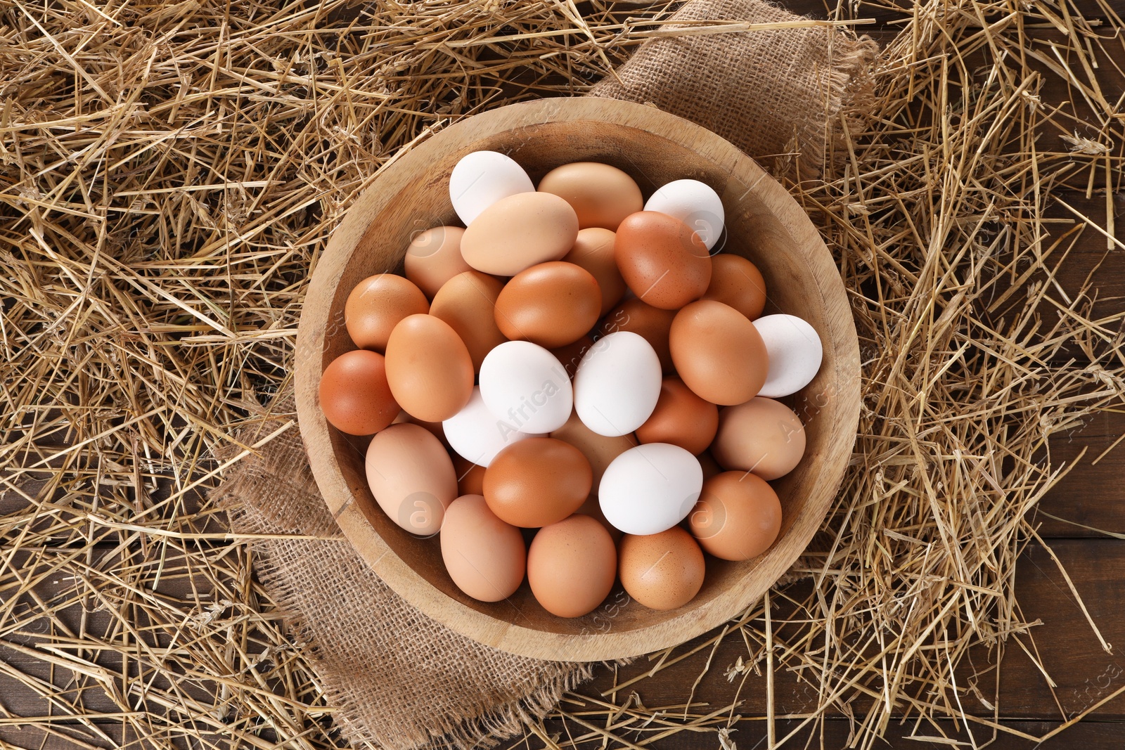 Photo of Fresh chicken eggs in bowl and dried straw on wooden table, top view