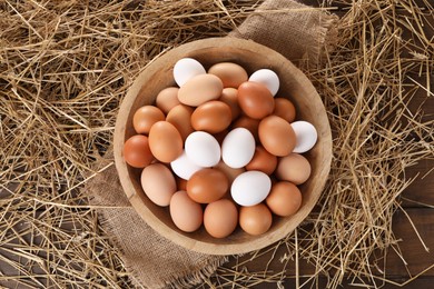 Fresh chicken eggs in bowl and dried straw on wooden table, top view