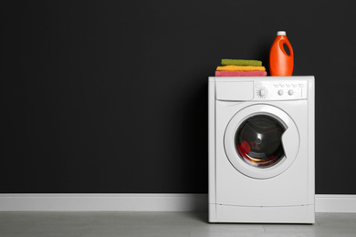 Photo of Modern washing machine with laundry, stack of towels and detergent near black wall. Space for text