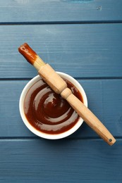 Tasty barbeque sauce in bowl and brush on blue wooden table, top view