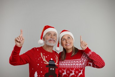 Senior couple in Christmas sweaters and Santa hats pointing at something on grey background. Space for text