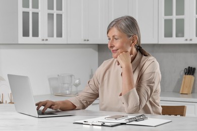 Photo of Beautiful senior woman using laptop at white marble table in kitchen, space for text