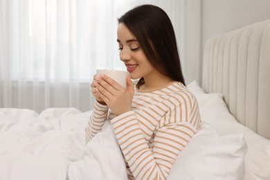 Beautiful young woman with cup of drink in bed at home