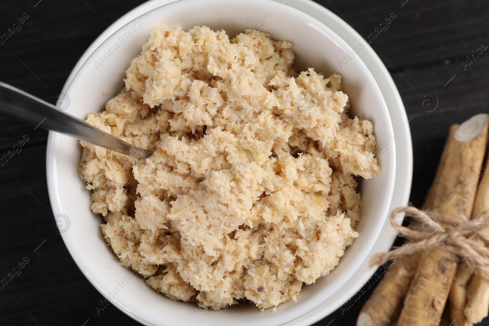 Photo of Bowl of tasty prepared horseradish, spoon and roots on black wooden table, flat lay