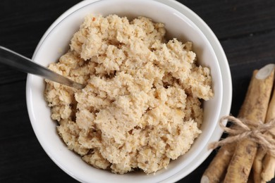 Photo of Bowl of tasty prepared horseradish, spoon and roots on black wooden table, flat lay