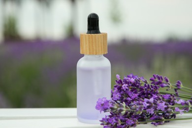 Bottle of essential oil and lavender flowers on white wooden table in field