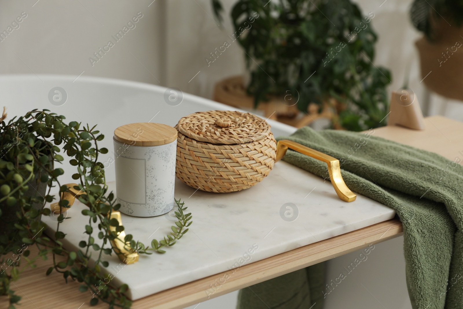 Photo of Tray with jar, wicker box, towel and green plant on bathtub indoors. Interior element