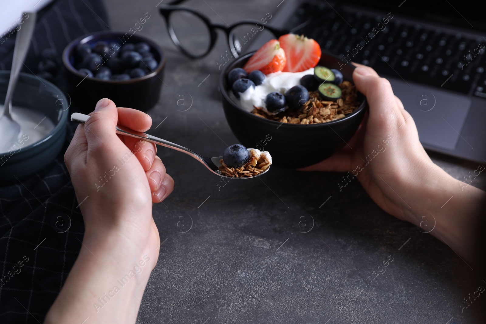 Photo of Woman eating tasty granola with yogurt and berries at workplace, closeup