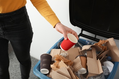 Photo of Young woman throwing coffee cup in trash bin indoors, closeup. Waste recycling