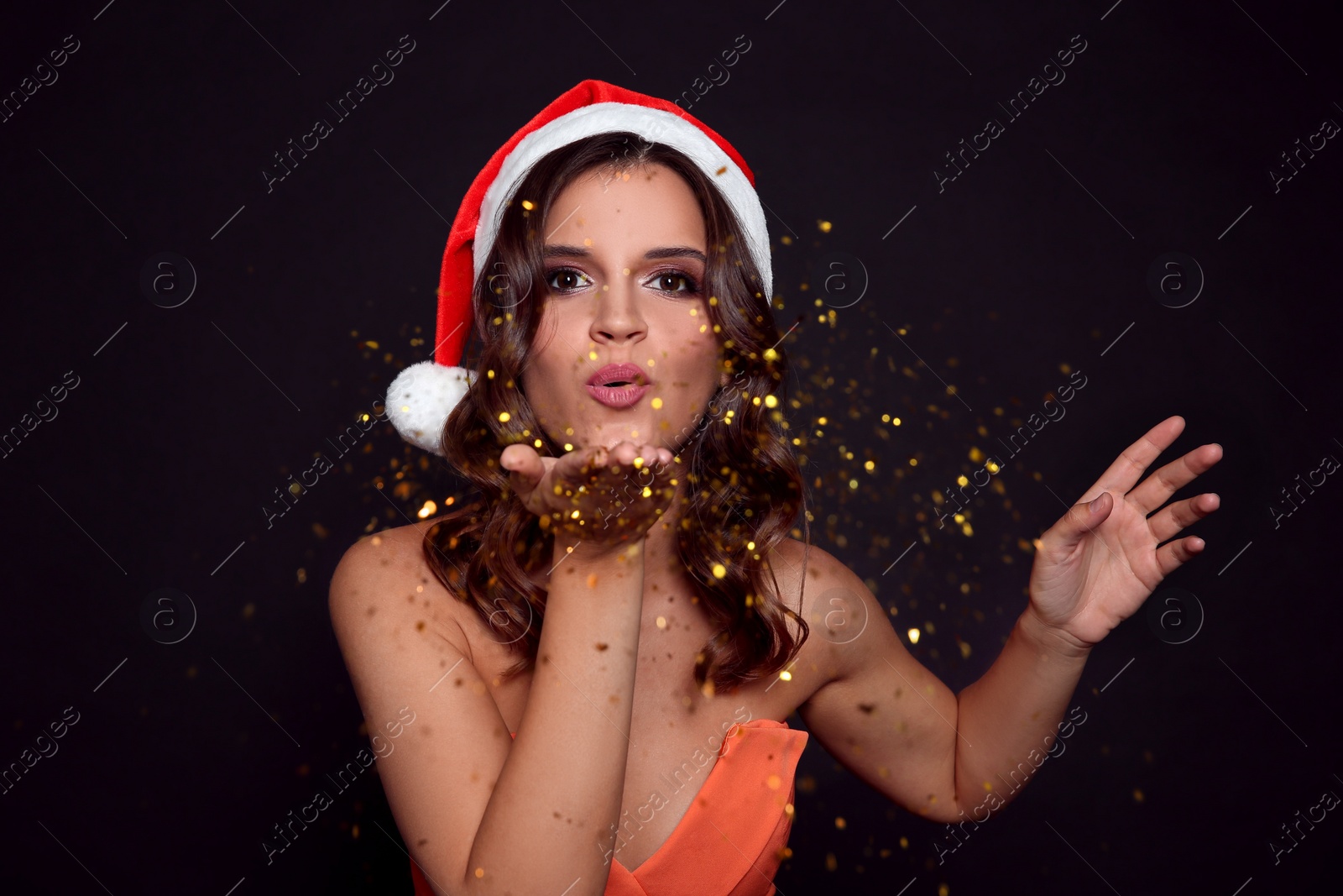 Photo of Beautiful woman in Santa hat blowing glitter on black background. Christmas party