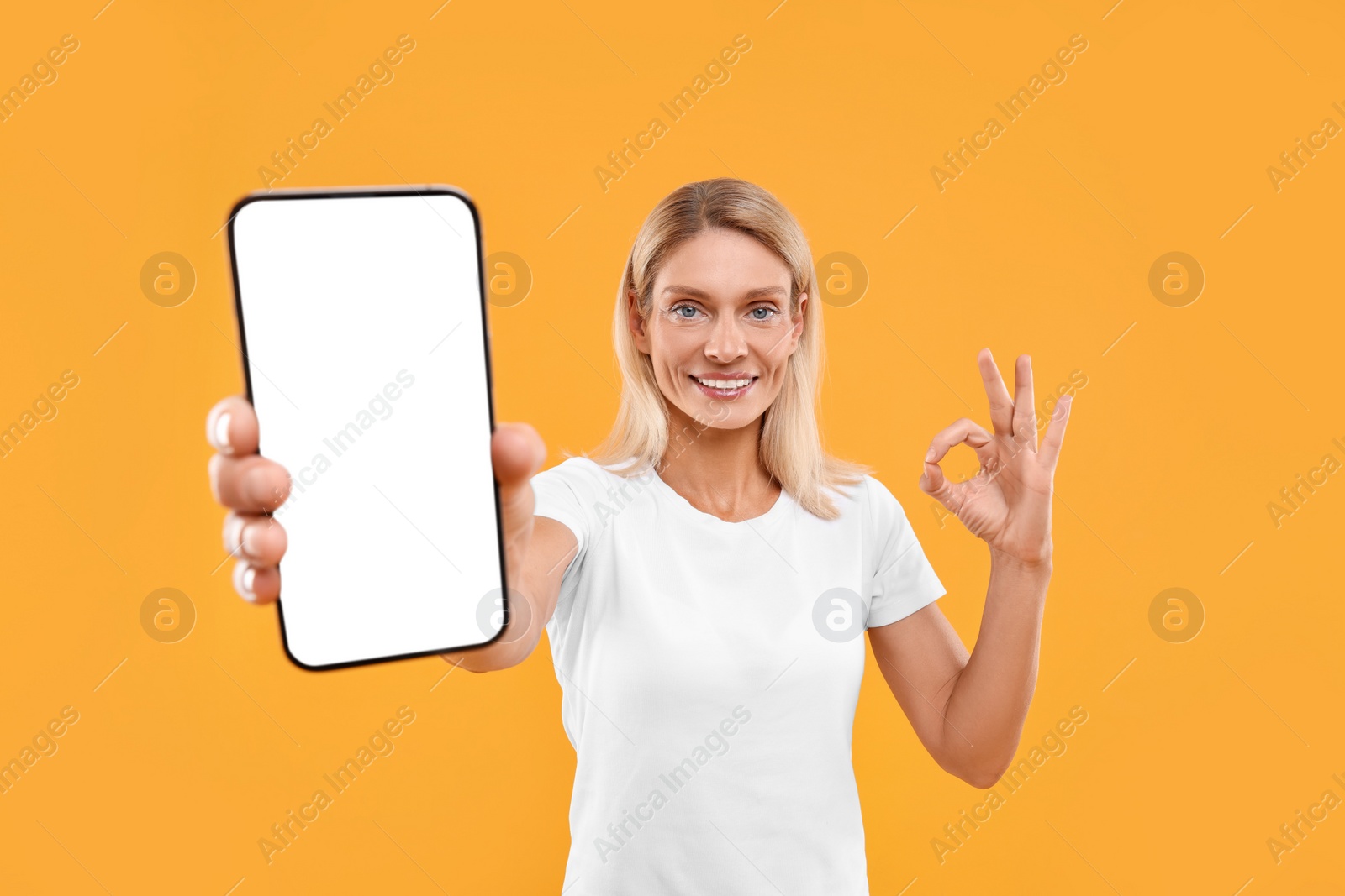 Photo of Happy woman holding smartphone with blank screen and showing OK gesture on orange background