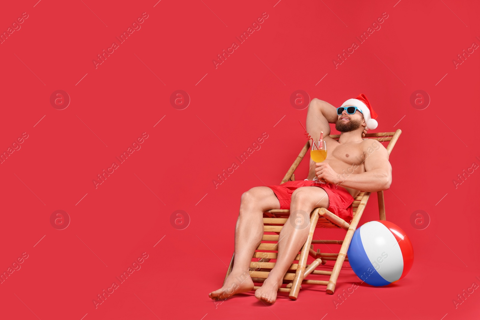 Photo of Muscular young man in Santa hat with cocktail, ball, sunglasses and deck chair on red background, space for text