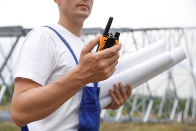 Professional engineer with plans and walkie talkie near high voltage tower construction outdoors, closeup. Installation of electrical substation