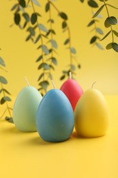 Photo of Colorful egg shaped candles and leaves on yellow background. Easter decor