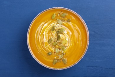 Photo of Bowl of delicious pumpkin soup with seeds and cream on blue wooden table, top view
