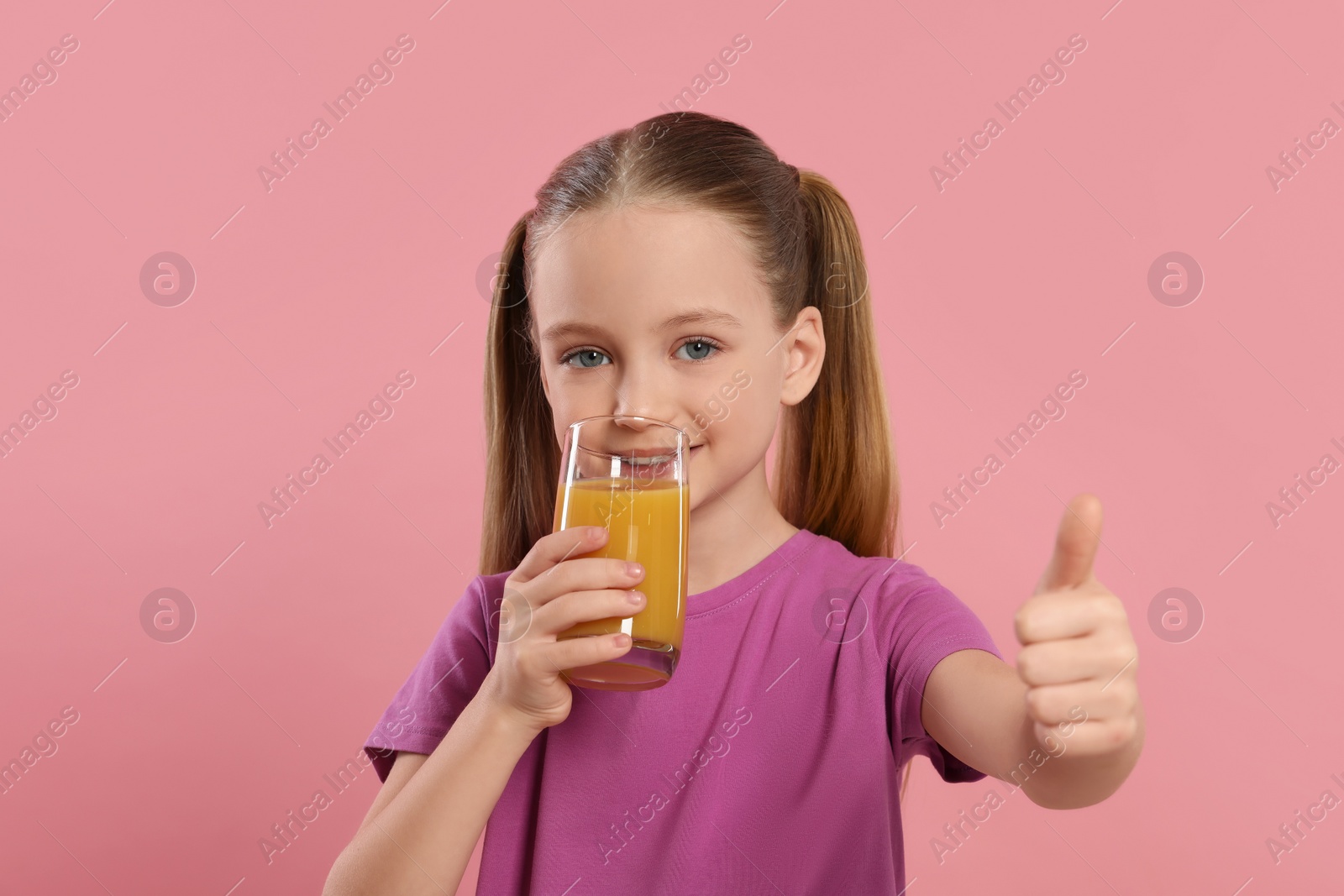 Photo of Cute little girl with glass of fresh juice showing thumbs up on pink background