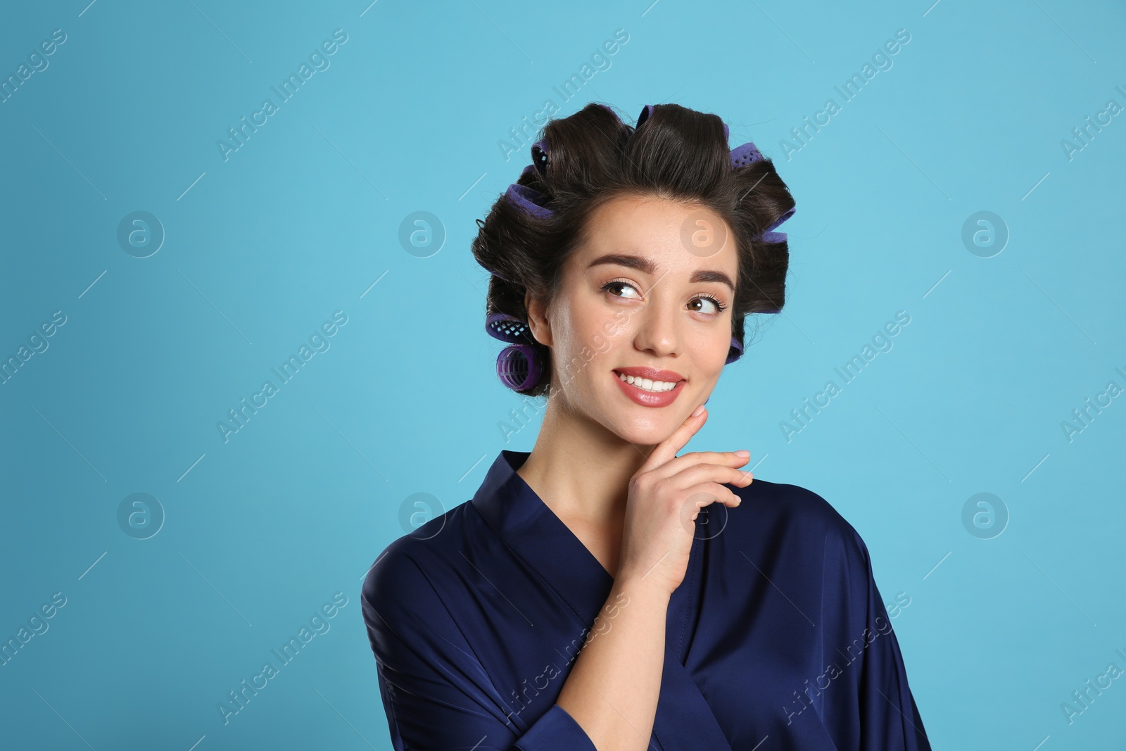 Photo of Happy young woman in silk bathrobe with hair curlers on light blue background, space for text