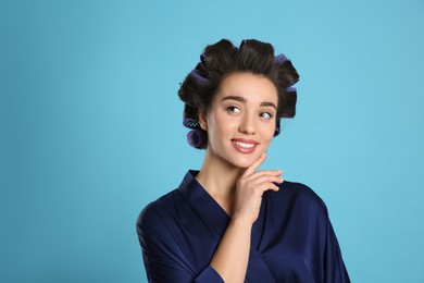 Happy young woman in silk bathrobe with hair curlers on light blue background, space for text