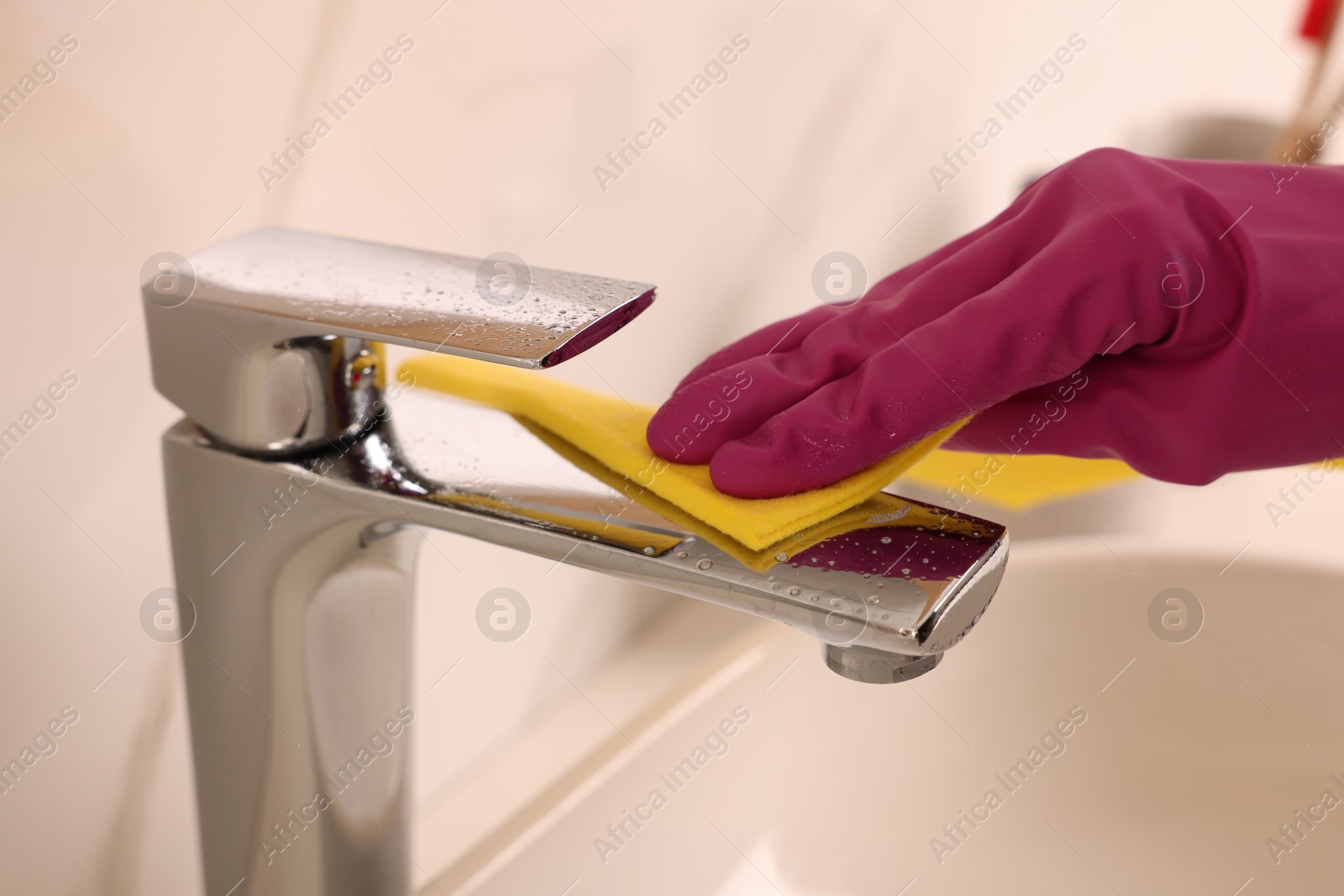 Photo of Woman in gloves cleaning faucet of bathroom sink with rag, closeup