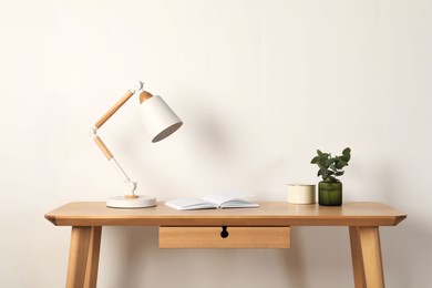 Photo of Stylish modern desk lamp, open book and plant on wooden table near white wall