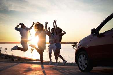 Photo of Happy friends jumping near car outdoors at sunset. Summer trip