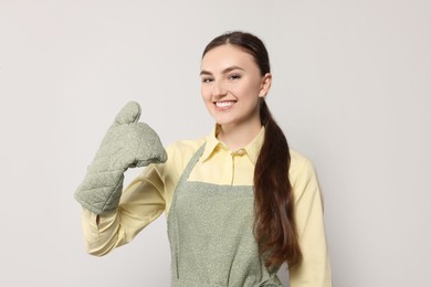 Photo of Beautiful young woman in clean apron with pattern and oven glove on light grey background