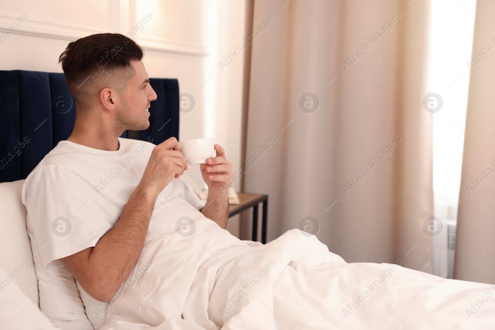 Photo of Handsome man drinking coffee on bed in hotel room