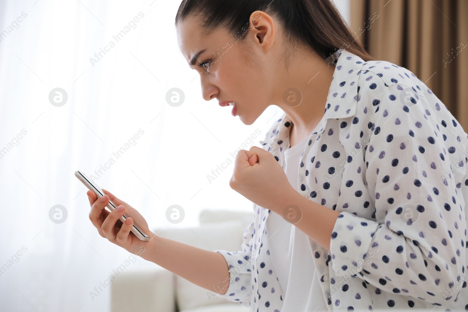 Photo of Emotional young woman with smartphone at home. Online hate concept