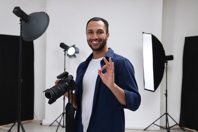 Photo of Young professional photographer with camera in modern photo studio showing OK gesture