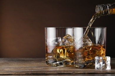 Photo of Pouring whiskey into glass with ice cubes at wooden table against brown background, space for text
