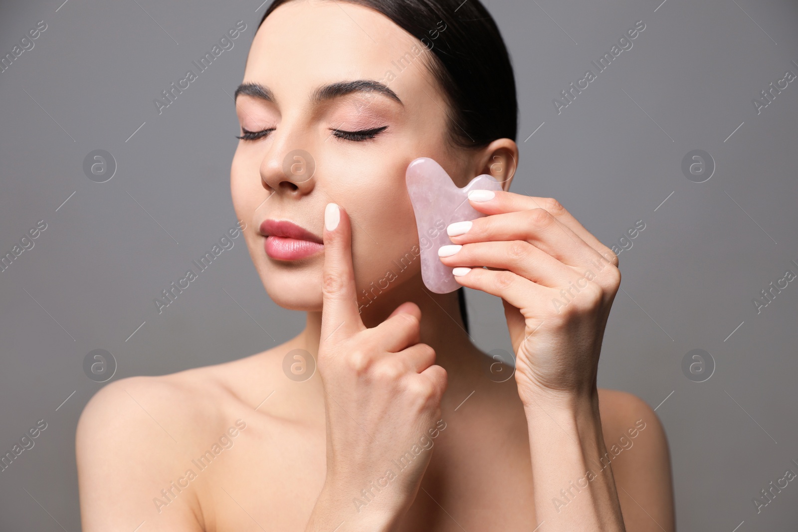 Photo of Beautiful young woman doing facial massage with gua sha tool on grey background, closeup