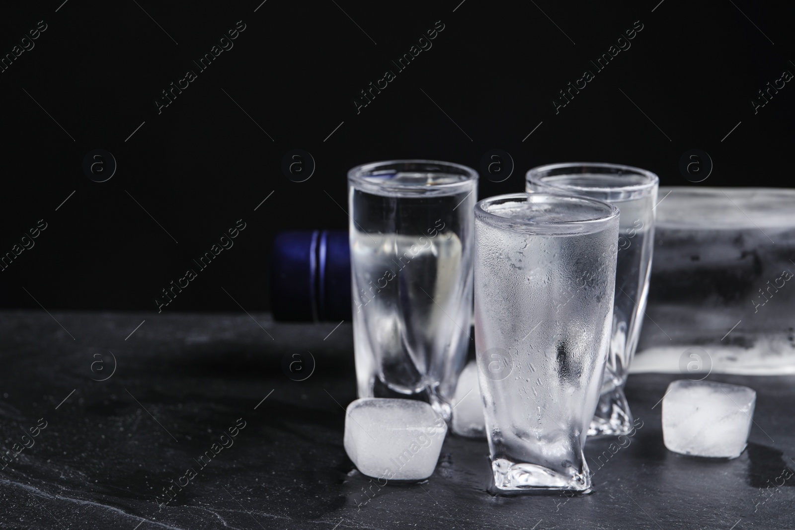 Photo of Bottle of vodka and shot glasses with ice on table against black background. Space for text