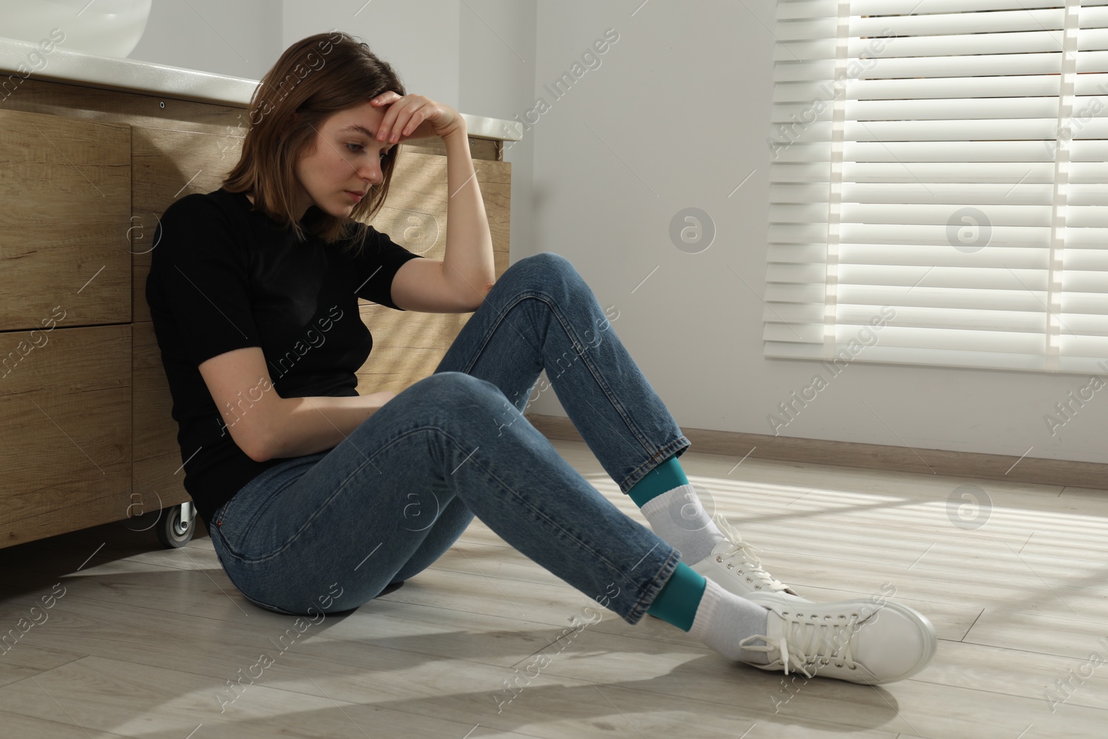 Photo of Sad young woman sitting on floor in bathroom, space for text