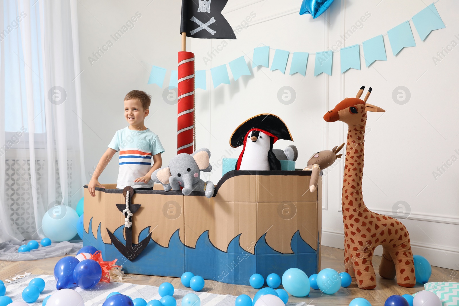 Photo of Cute little boy playing with pirate cardboard ship and toys at home. Child's room interior