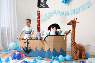 Cute little boy playing with pirate cardboard ship and toys at home. Child's room interior