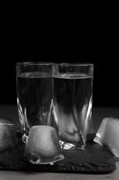 Photo of Shot glasses with vodka and ice cubes on grey tale against black background, closeup
