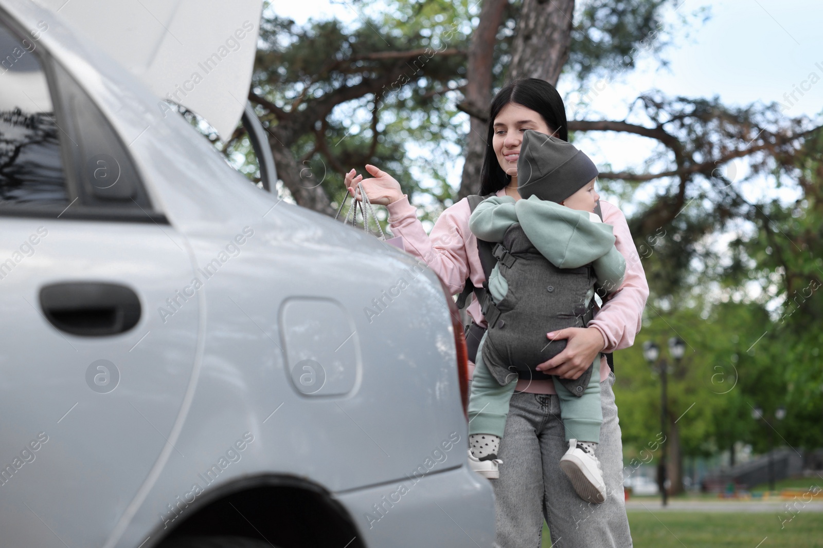 Photo of Mother holding her child in sling (baby carrier) while putting shopping bags into car trunk outdoors