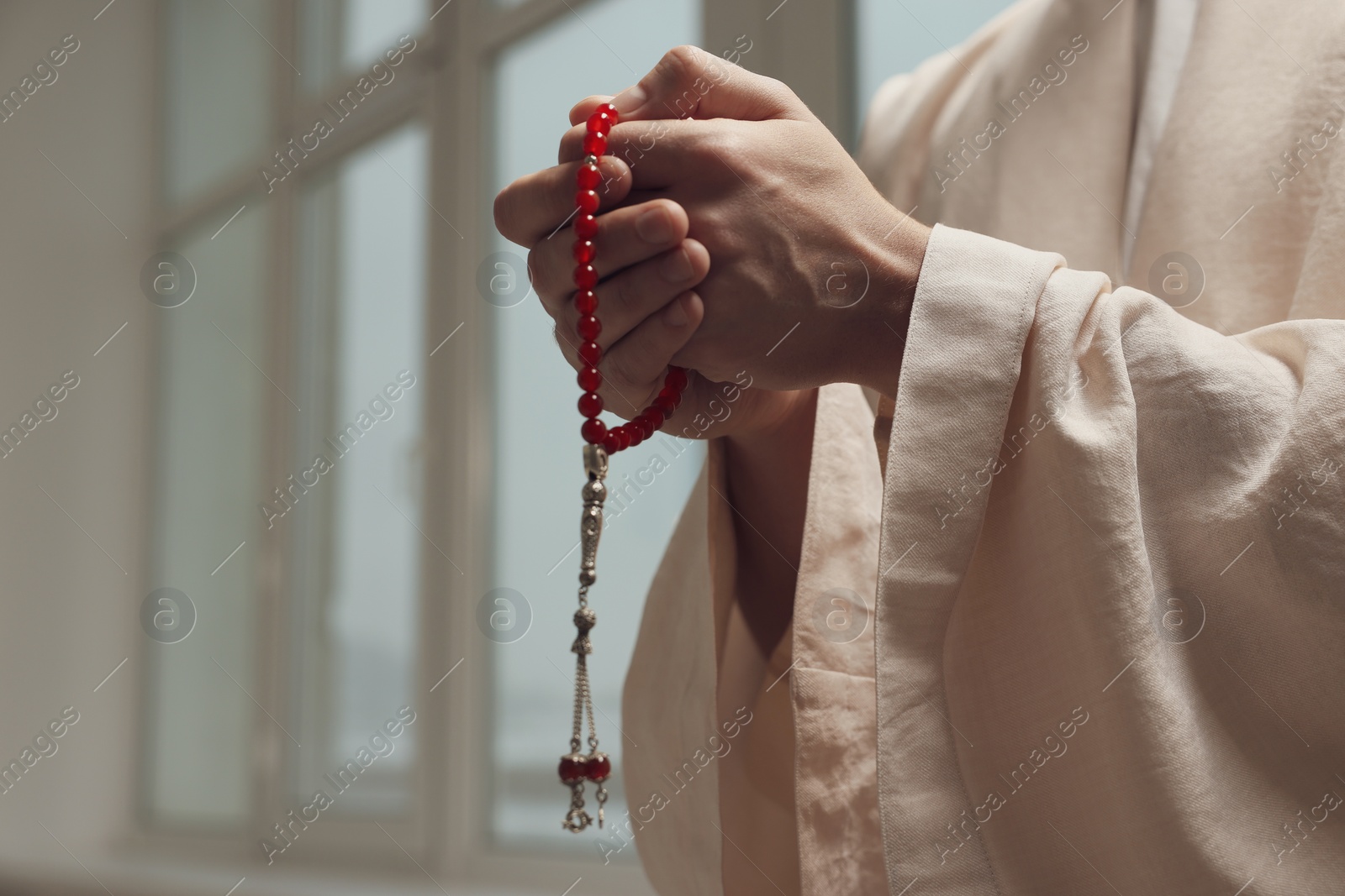 Photo of Muslim man with misbaha praying near window indoors, closeup. Space for text