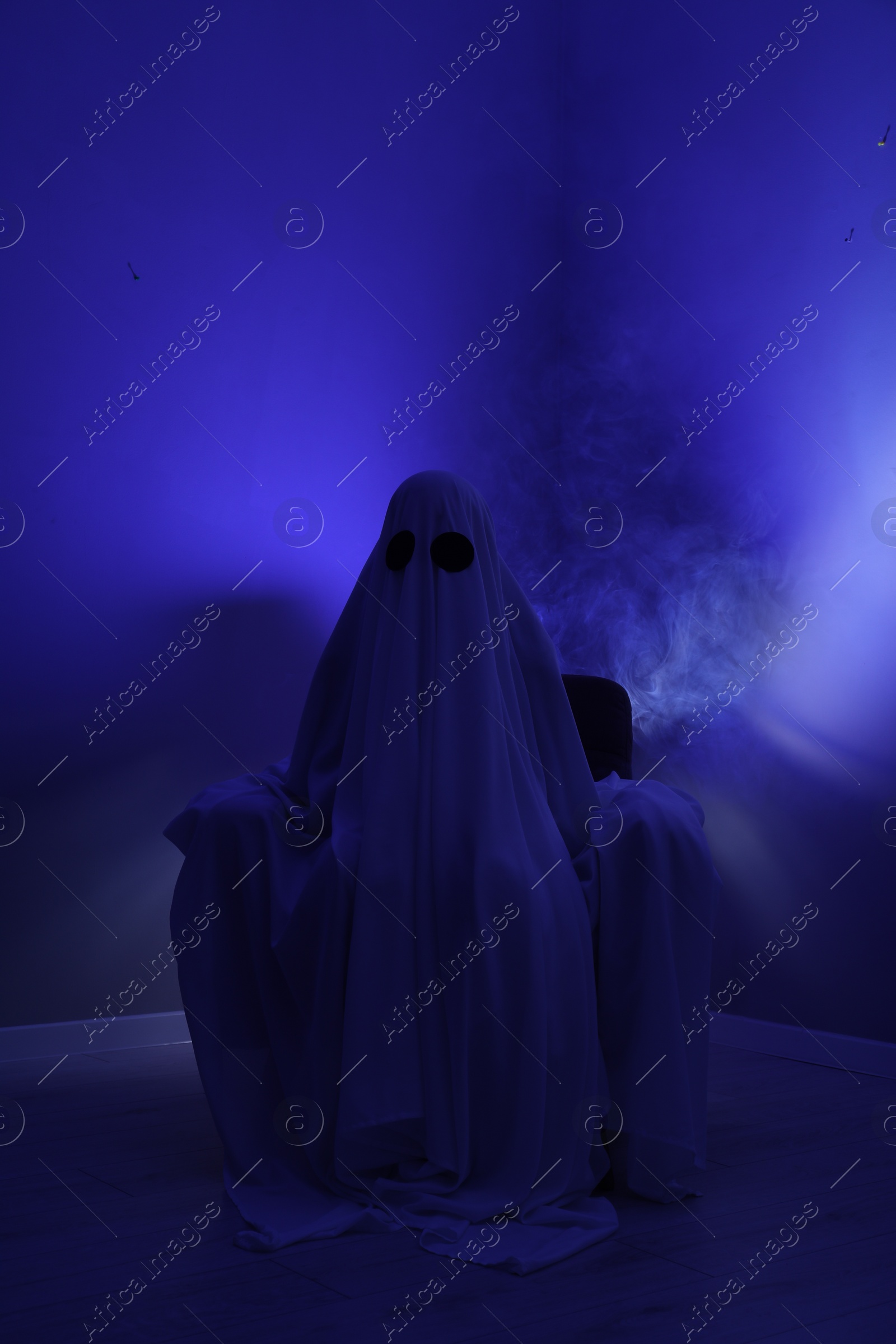 Photo of Creepy ghost. Woman covered with sheet sitting in armchair in blue light