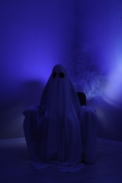 Creepy ghost. Woman covered with sheet sitting in armchair in blue light