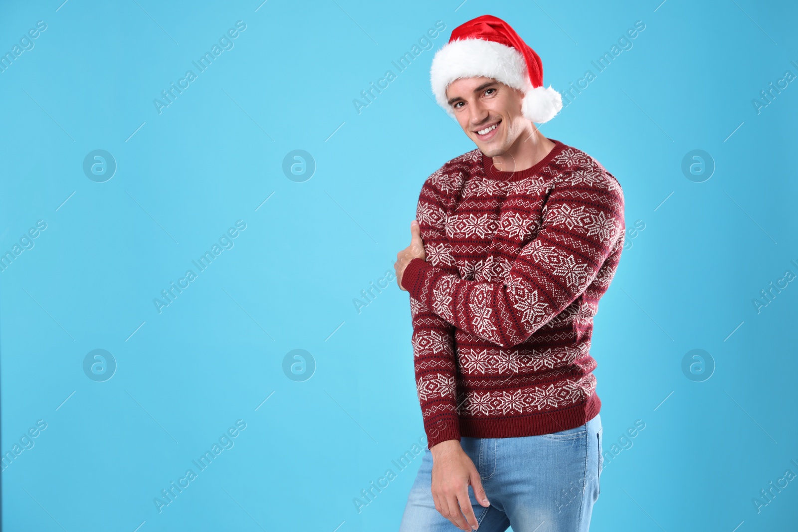 Photo of Portrait of young man in Christmas sweater and Santa hat on light blue background. Space for text