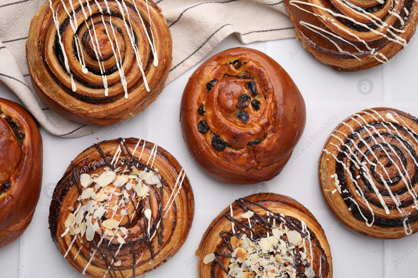 Photo of Delicious rolls with raisins, nuts and toppings on white tiled table, flat lay. Sweet buns