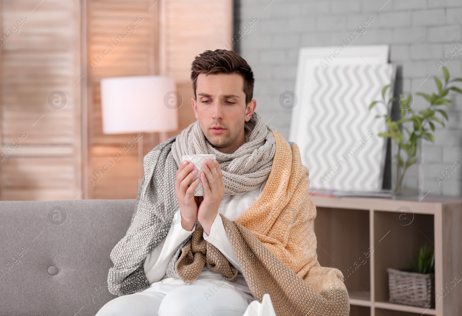 Photo of Sick young man with cup of hot drink suffering from cold at home