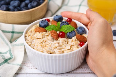 Woman holding bowl of oatmeal with berries, almonds and mint at white table, closeup