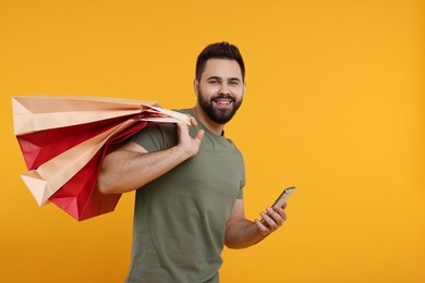 Smiling man with many paper shopping bags and smartphone on orange background. Space for text