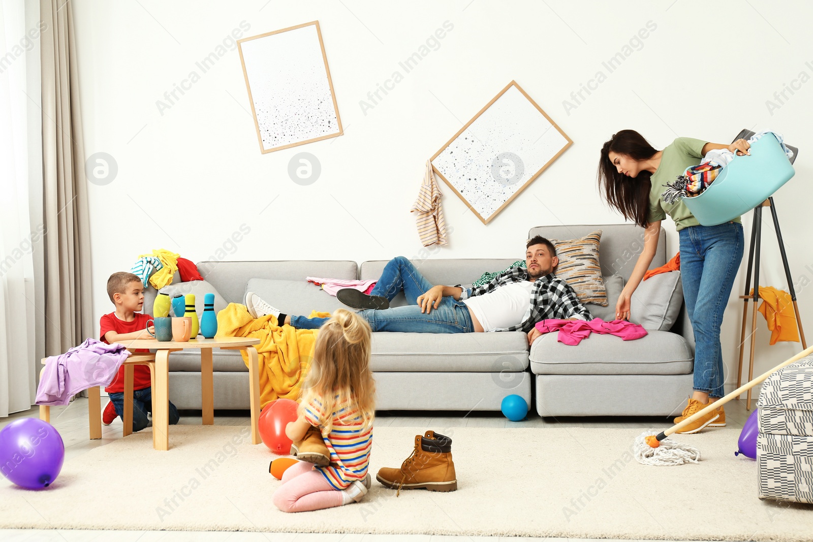 Photo of Exhausted mother trying to clean up mess made by children and lazy father in room