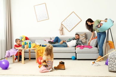 Photo of Exhausted mother trying to clean up mess made by children and lazy father in room