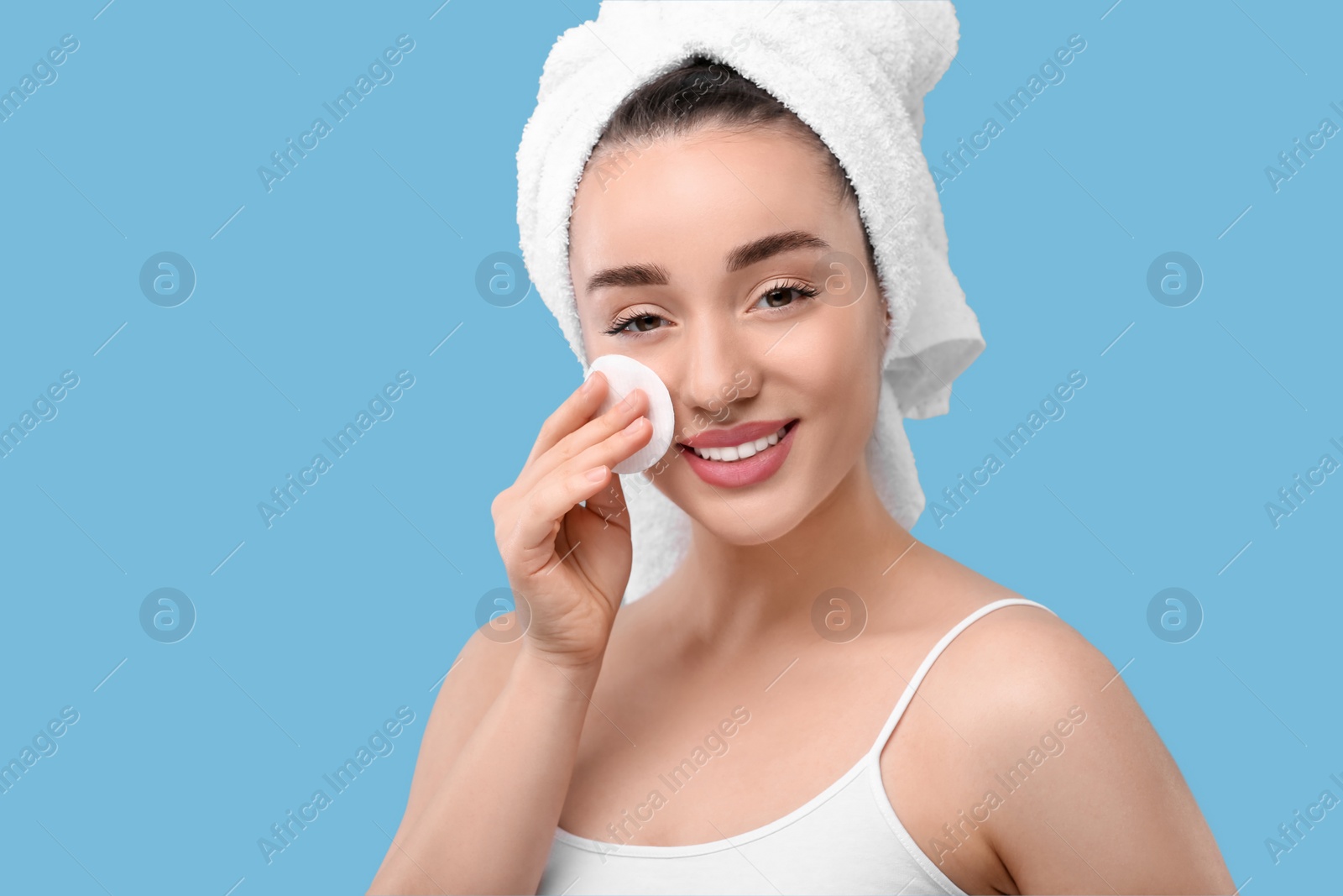 Photo of Beautiful woman in terry towel removing makeup with cotton pad on light blue background